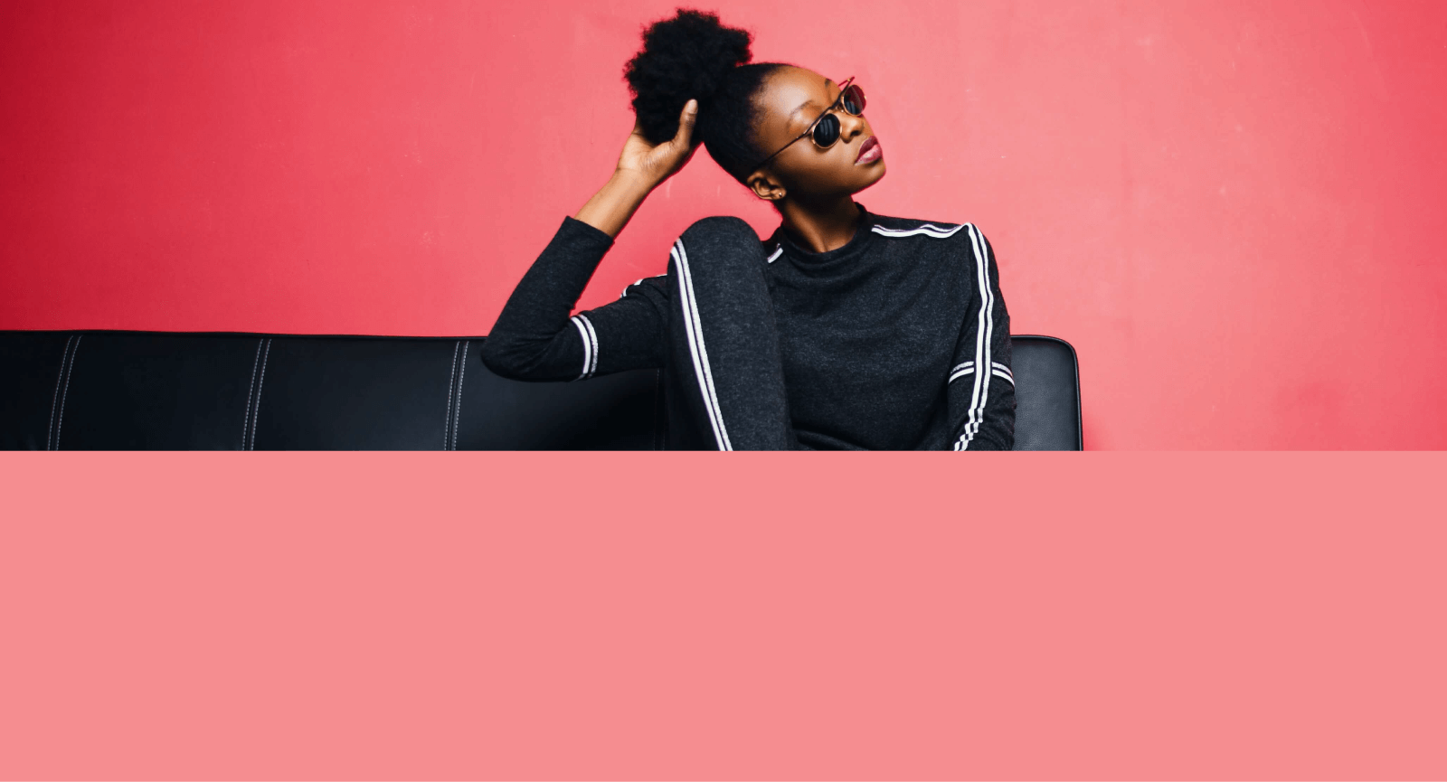 Modelo feminina utilizando óculos escuros e vestindo conjunto esportivo de blusa e calça na cor preta, sentada em um sofá preto em um fundo rosa